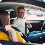 A husband puts on his seatbelt and smiles at his wife from the driver’s seat of their car. 